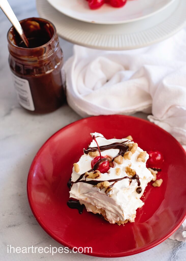 A single slice of Banana Split Lasagna served on a red plate next to a jar of fudge ice cream topping.