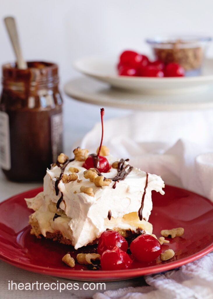A single slice of Banana Split Lasagna - layers creamy banana pudding, whipped topping, fudge, and a graham cracker crust, topped with walnuts, fudge drizzle, and maraschino cherries.