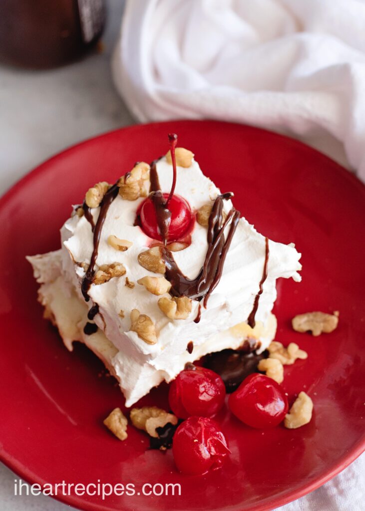 A single slice of banana split lasagna served on a red plate. The creamy dessert is topped with a layer of whipped topping, walnut pieces, cherries, and a drizzle of fudge topping.