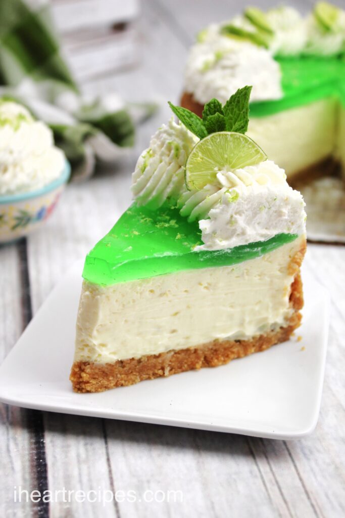 A single slice of key lime Jello cheesecake on a small white plate. The slice of cake has a graham cracker crust, thick layer of key lime cheesecake, a thin layer of lime green Jello, and is topped with whipped cream, a lime wedge, and lime zest.