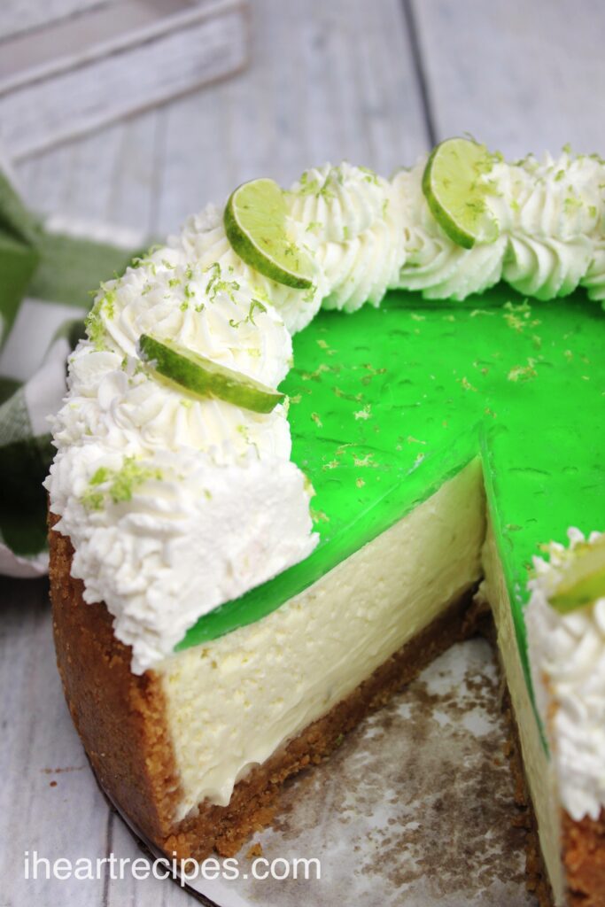 A whole key lime Jello cheesecake that's topped with a layer of lime green Jello, whipped cream, and lime zest. A single slice is cut from the cake, revealing the layers of cheesecake and crust at the bottom.