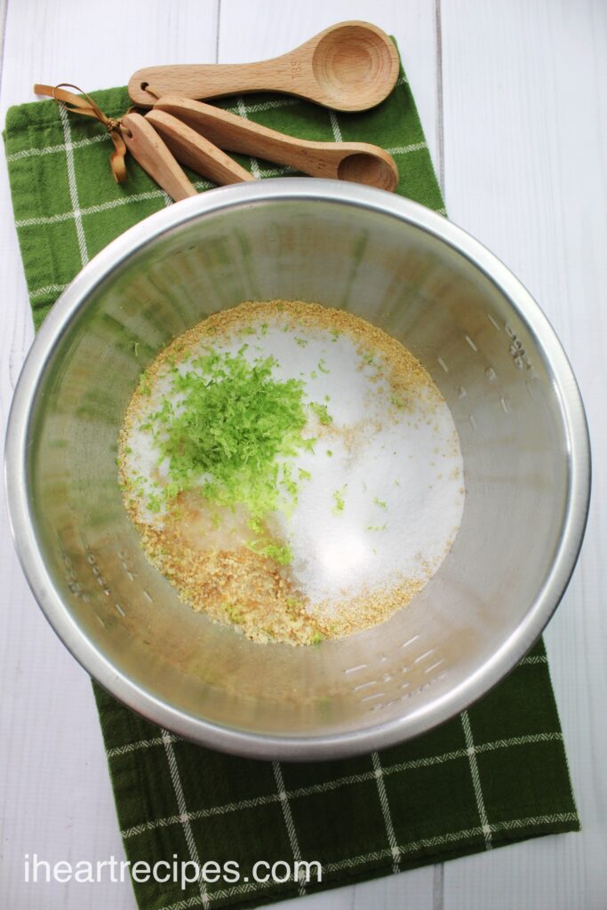 An overhead image of a metal bowl filled with ingredients to make the graham cracker crust of a key lime Jello cheesecake - crushed graham crackers, white sugar, butter, and lime zest.