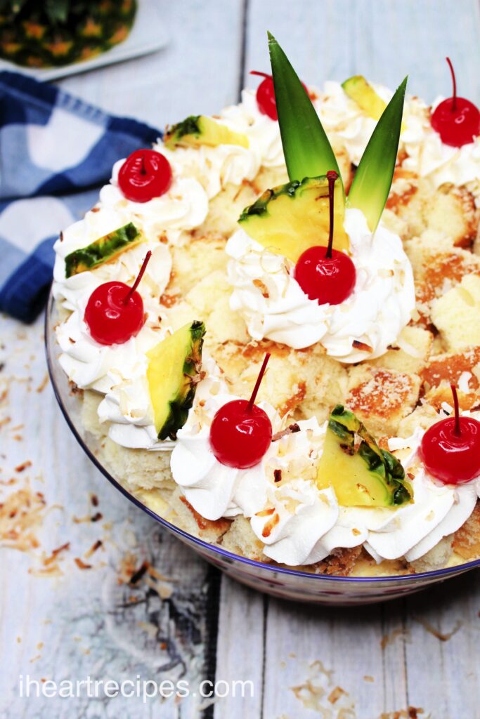 A close up image of the top of a pina colada cake trifle, topped with dollops of whipped cream, pineapple chunks, and maraschino cherries.