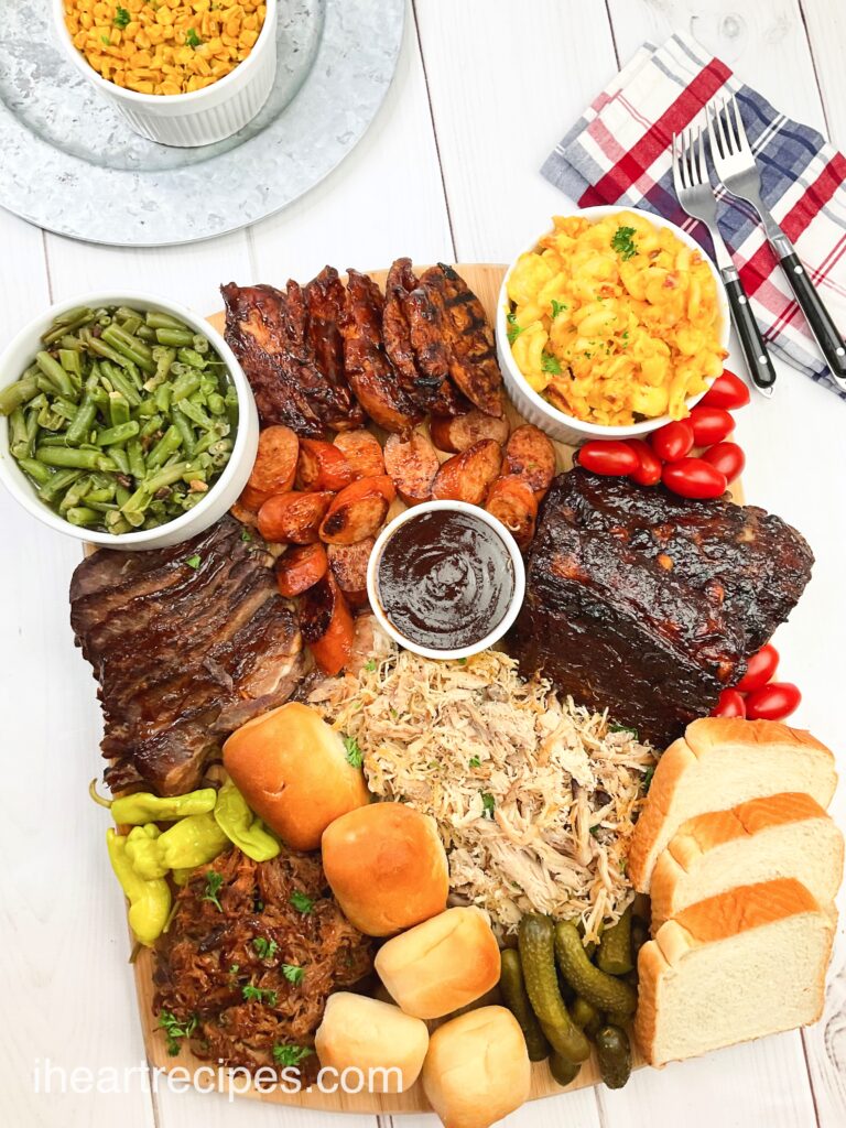 An overhead shot of a filled leftover BBQ charcuterie board, packed with BBQ chicken, sausage, ribs, and brisket, bread, rolls and sauces. Bowls of sides - green beans and mac and cheese - fill out the extra spaces.