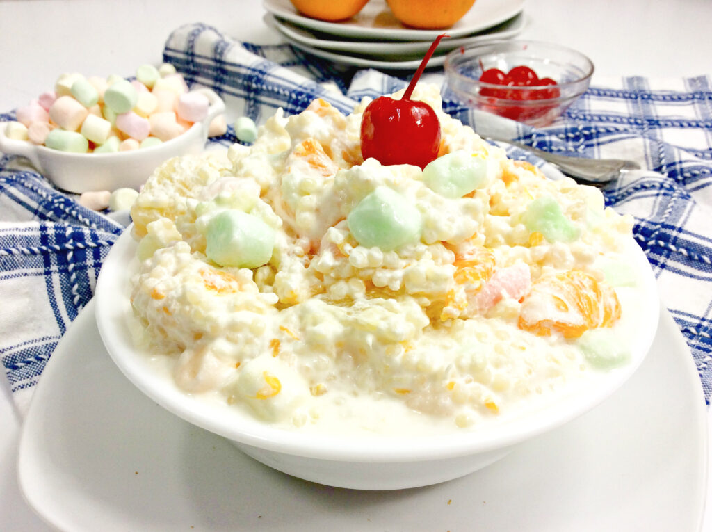 A bowl of frog eye salad made with acini de pepe pasta mixed with a whipped topping and mixed fruit, topped with marshmallows and a single cherry.