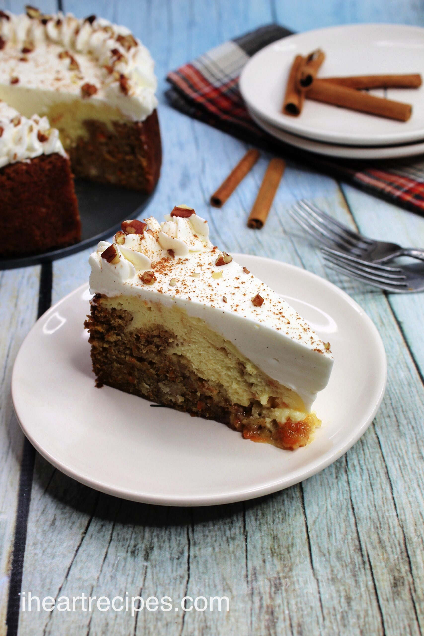 A slice of carrot cake cheesecake is served on a white plate. In the background, the whole cheesecake is set on a springform pan platter.