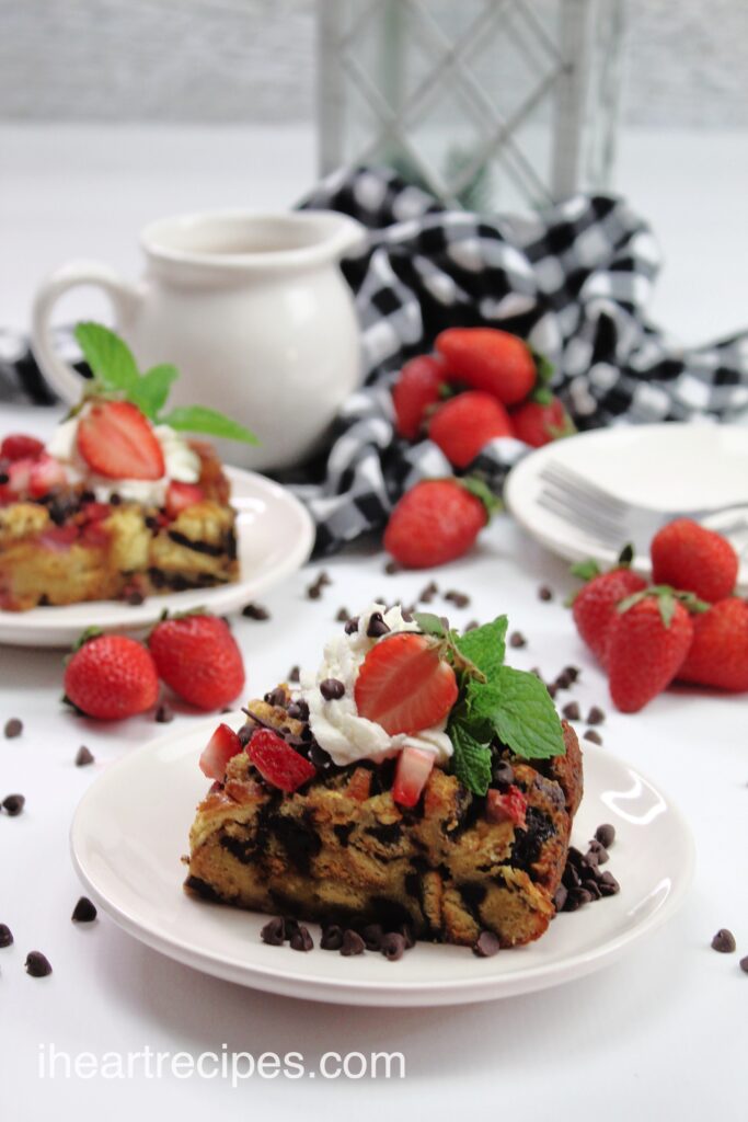 A slice of Chocolate Strawberry French Toast Casserole served on a round white plate. There is another serving, a white pitcher, a black and white checked tablecloth and fresh whole strawberries seen in the background. 