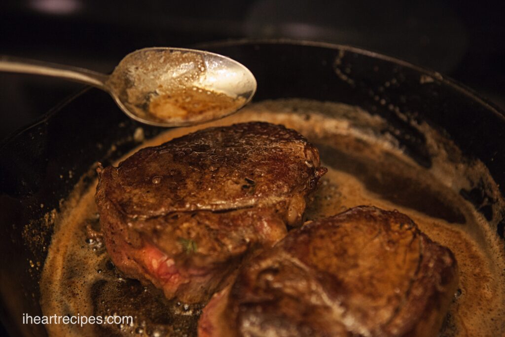 Two seared filet mignon in a cast iron skillet. A silver spoon is drizzling golden herb butter over the steaks.