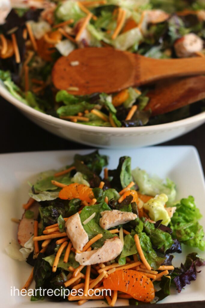 A serving of Fresh Mandarin Orange Salad is displayed on a small white square plate. A large salad bowl filled with salad and a pair of wooden salad spoons is seen in the background. 