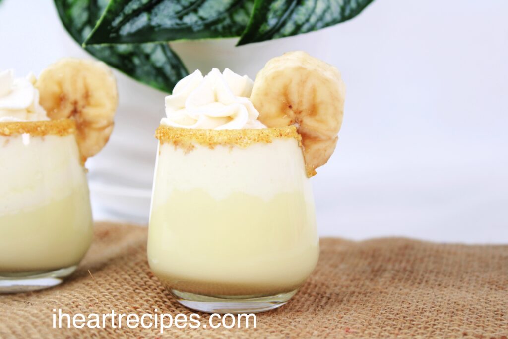 Two clear shot glasses filled with banana cream pie shots, rimmed with crushed graham crackers and a banana slice. They're set on a burlap tablecloth. There are dark green plant leaves visible in the background.