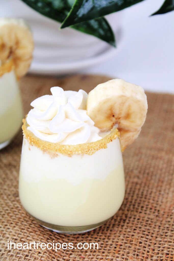 A shot of banana cream pie topped with a dollop of whipped cream and rimmed with graham crackers and a slice of banana. 