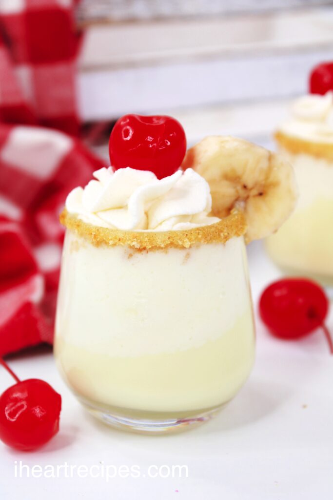A closeup of a clear shot glass filled with smooth banana cream, topped with whipped cream and a cherry. The rim is covered in graham cracker crumbs and a slice of banana. Maraschino cherries lay nearby. 