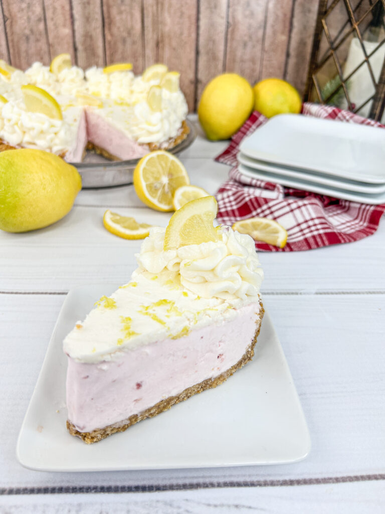 A creamy wedge of Strawberry Lemonade Dream Pie is served on a square white plate. The wedge is topped with a dollop of whipped cream and a lemon slice. 