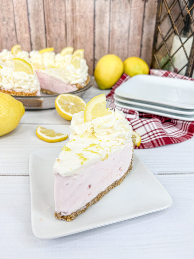 A slice of Strawberry Lemonade Dream Pie topped with a dollop of whipped cream and a lemon slice served on a square white plate. The remaining pie is seen in the background. Bright yellow lemons, white plates and a red and white checked cloth napkin lay nearby.  