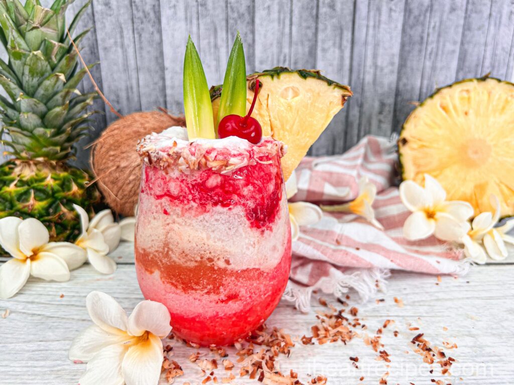 Red, orange and cream layers of this island crush frozen margarita show through a clear tumbler. It's garnished with a coconut rim, pineapple leaves and cherry. Tropical fruit, flowers and toasted coconut lay nearby. 