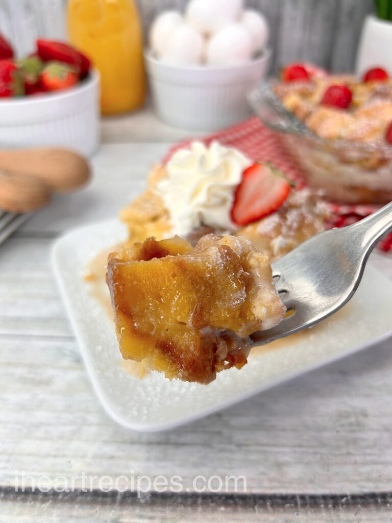 A silver fork holds a single bite of French Toast casserole. In the background is a slice the French toast bake on a plate, and a full casserole dish.