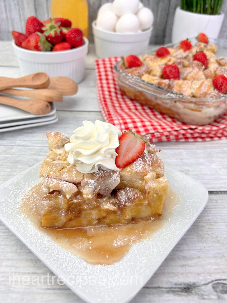 A serving of French toast casserole drenched in sweet syrup and topped with a dollop of whipped cream and a strawberry slice.