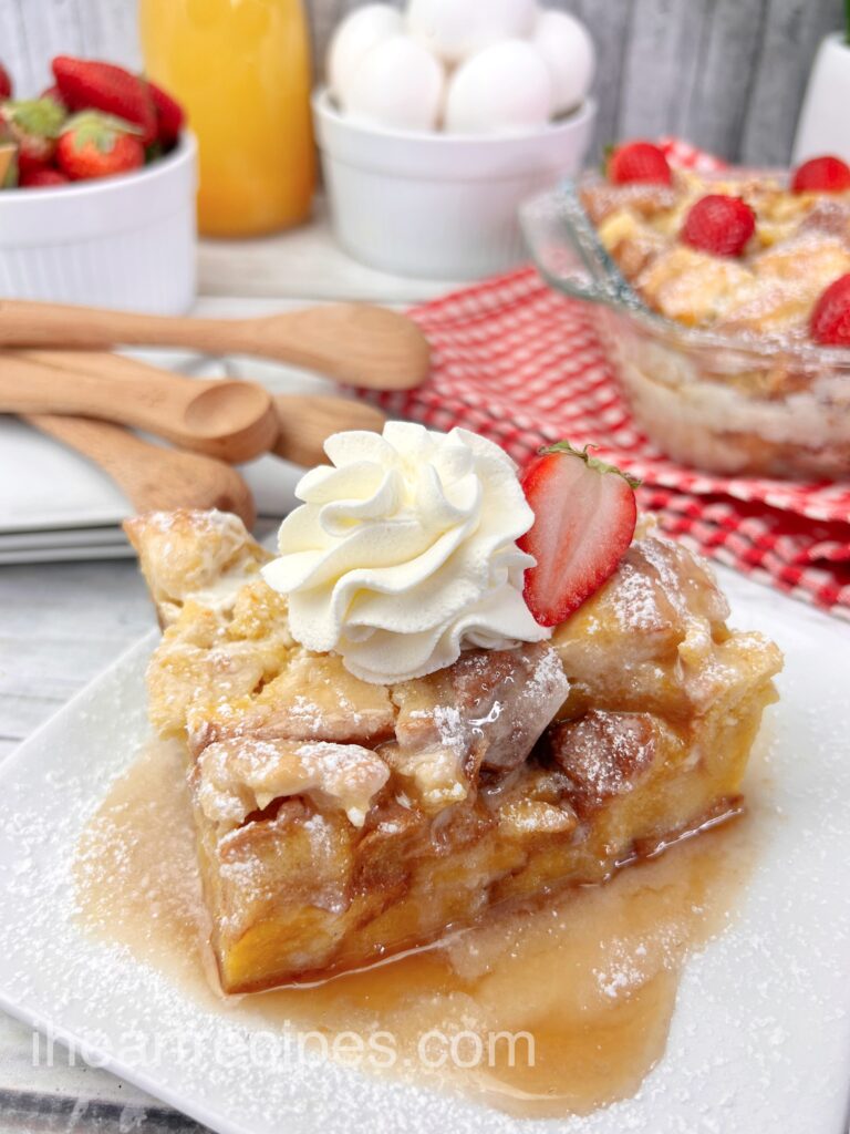 A slice of homemade French toast casserole served on a plate, topped with a strawberry slice, whipped cream, and syrup.