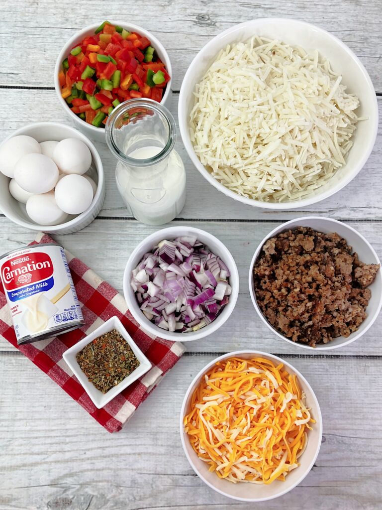 An overhead image of the ingredients needed for hash brown casserole. Each ingredient is in it's own dish -- frozen hash browns, onions and peppers, sausage, cheese, eggs, milk, evaporated milk, and seasonings.