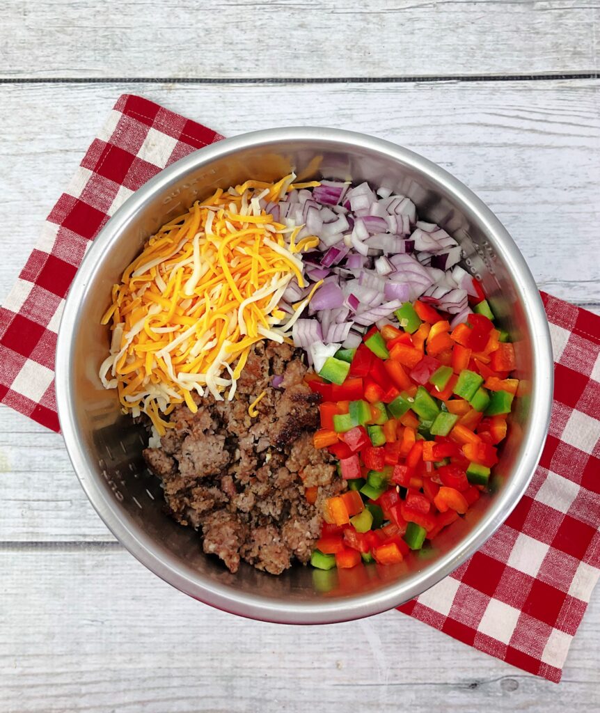 Shredded cheese, cooked ground sausage, onions, and red and green bell peppers mixed together in a metal mixing bowl.