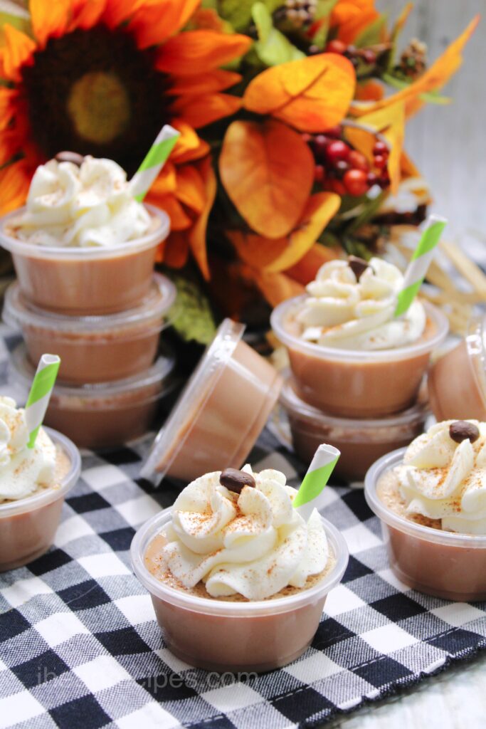 Stacks of pumpkin latte Jello shots arranged in front of a festive Fall flower arrangement. Each Jello shot is topped with whipped cream and an espresso bean.