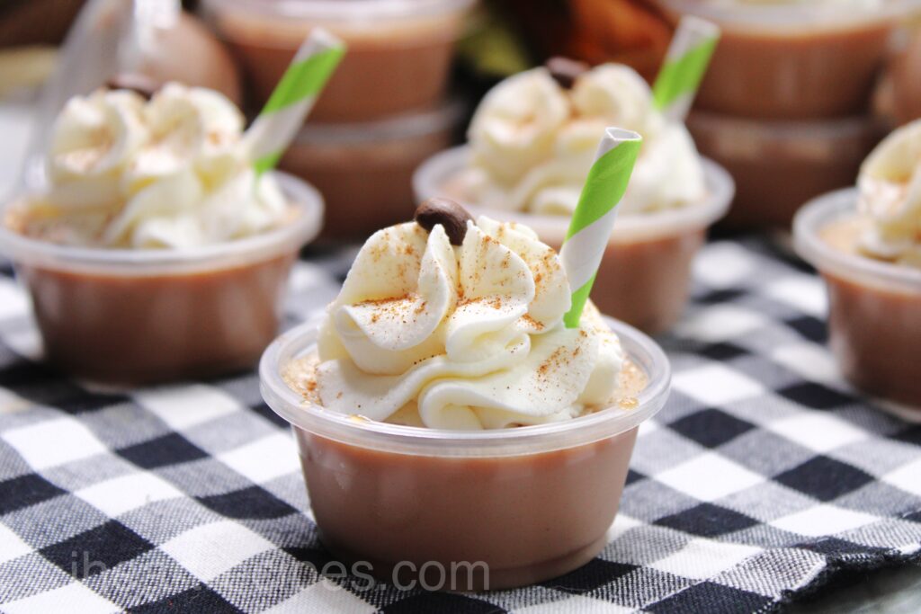 Pumpkin latte Jello shots lined up on a table, topped with whipped cream and a sprinkle of Pumpkin spice.