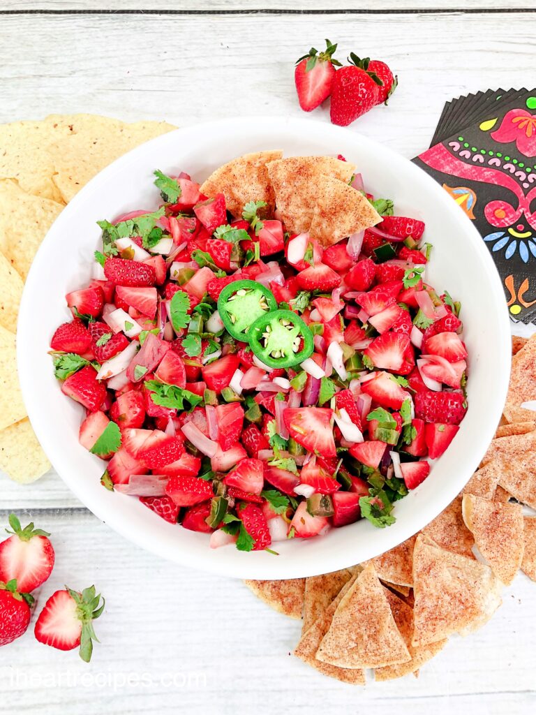 A big white serving bowl filled with fresh strawberry and jalapeño pico. Colorful napkins and tortilla chips lay nearby. 
