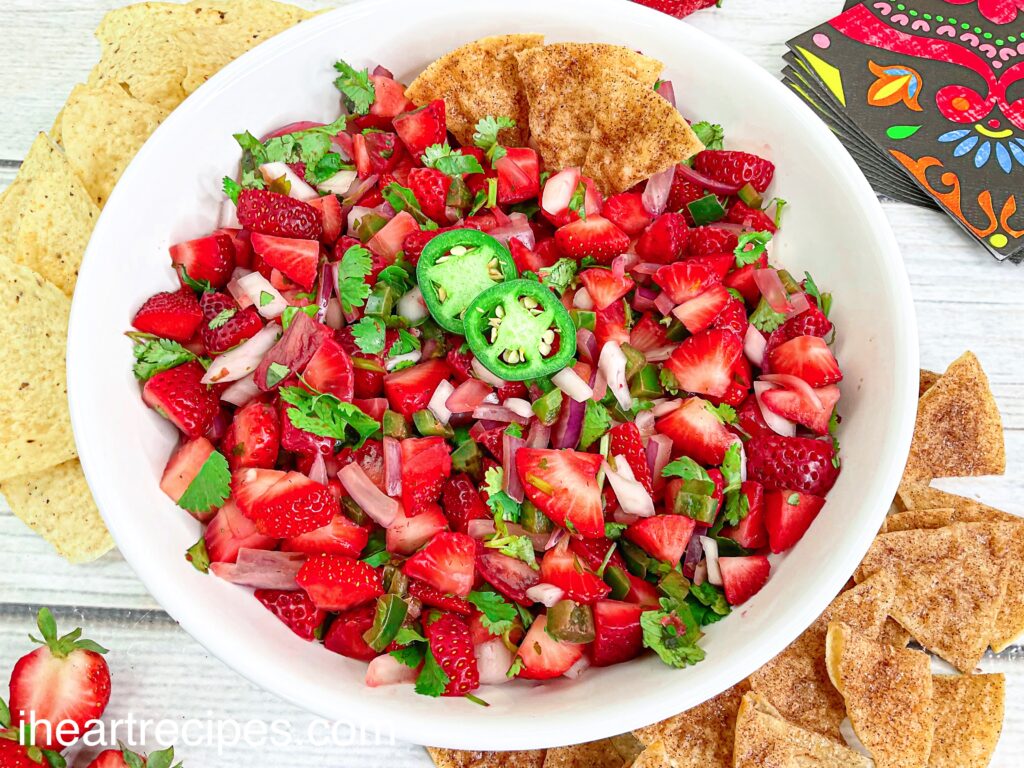 Vibrant reds and greens showoff in the strawberry and jalapeño pico served in a round white serving bowl. Plain and cinnamon tortilla chips surround the bowl.