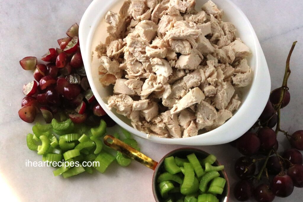 Diced chicken in a white bowl, sliced celery and quartered red grapes on a marble cutting board. 