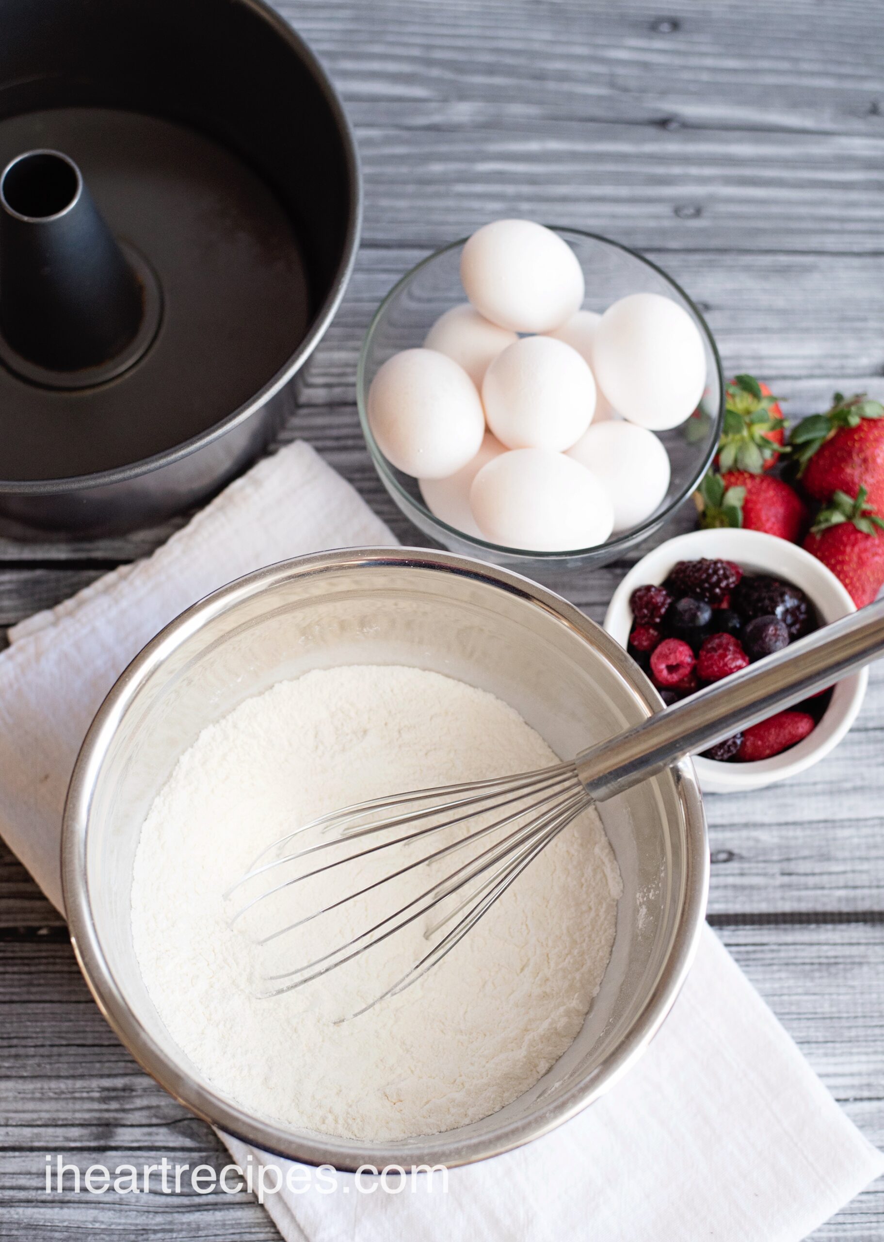 An overhead image of some of the ingredients needed to make homemade angel food cake -- eggs and a flour mixture in a bowl with a whisk. 