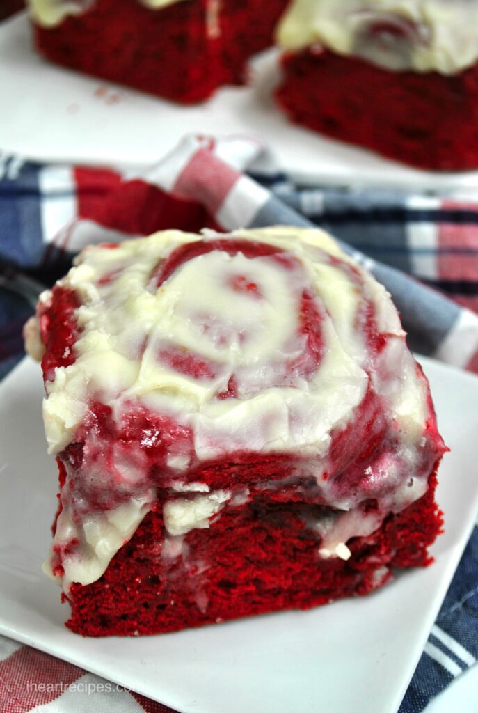 Red velvet cake mix cinnamon rolls topped with cream cheese frosting set served on square white plates.