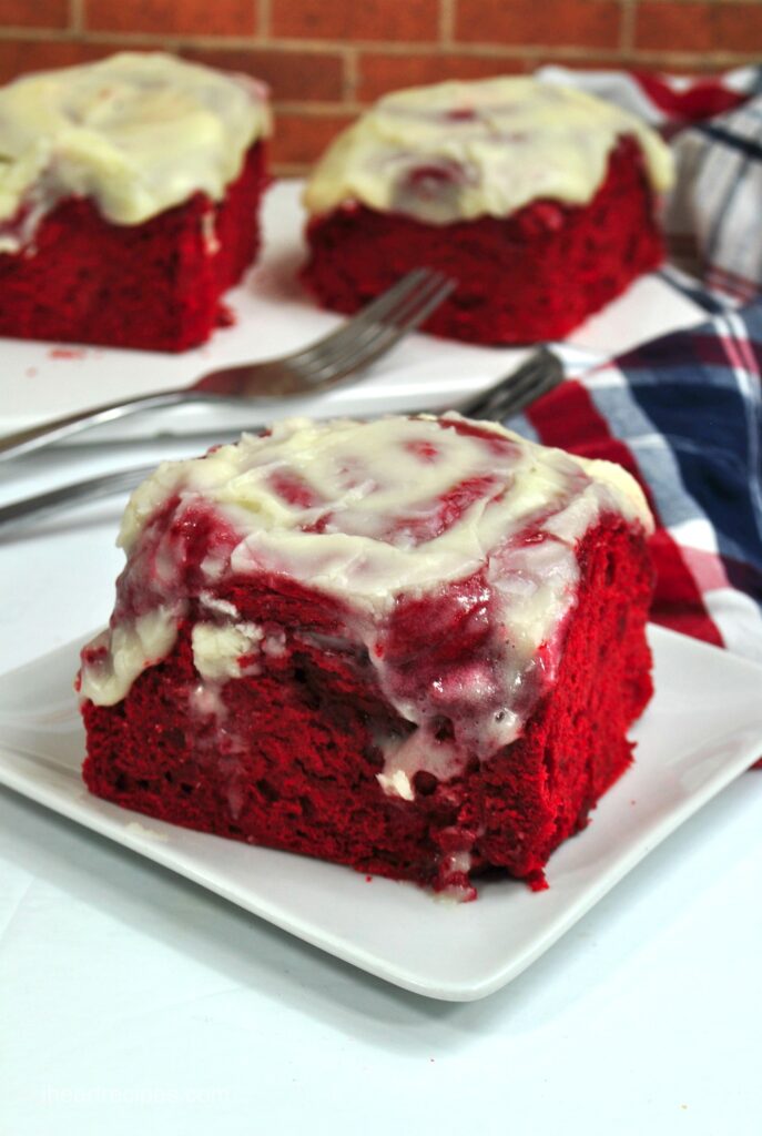 A close up of a red velvet cinnamon roll topped with cream cheese frosting melting down the side. 