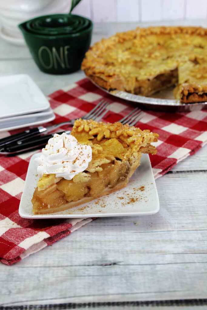 A slice of homemade apple pie cut from a whole pie, served with a dollop of whipped cream and a dusting of cinnamon.