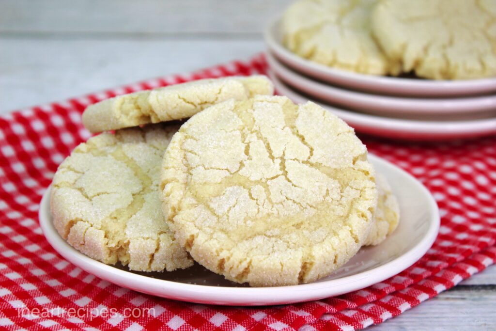 A plateful of crispy, chewy traditional sugar cookies lay on a red and white checkered tablecloth. 