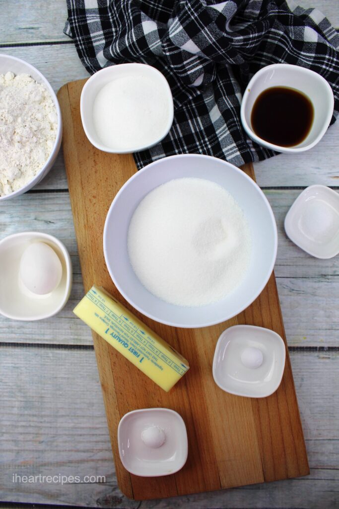 The ingredients for traditional sugar cookies in white bowls set out on a rustic wooden table.