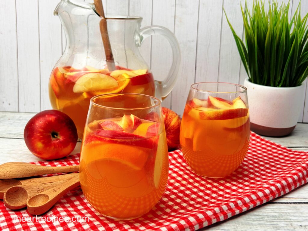 Two clear glasses filled with sliced apples and oranges and golden Apple Pie Sangria. A pitcher and apples are in the background. 