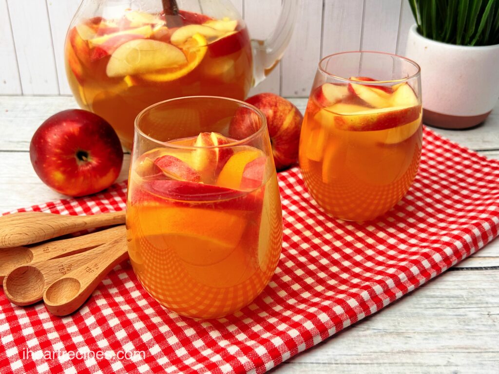 Two clear glasses and a clear glass pitcher filled with golden Apple Pie Sangria and sliced apples and oranges. Wooden measuring spoons and apples lay nearby.