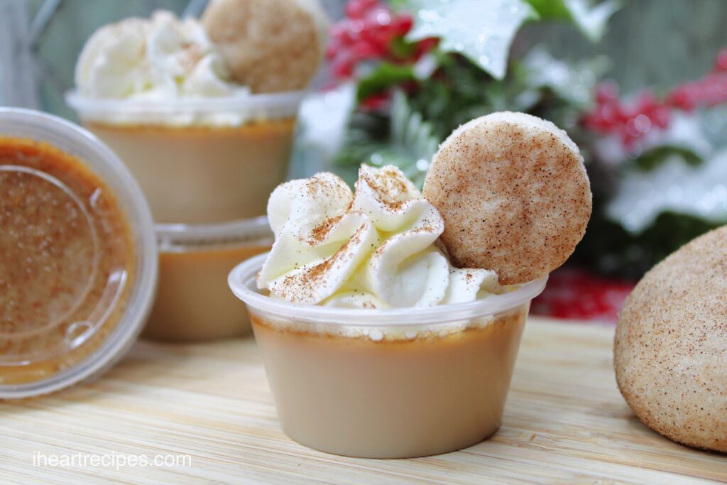 Personal servings of Snickerdoodle Jello Shots garnished with whipped cream and miniature Snickerdoodle cookies. Festive decorations are seen in the background.