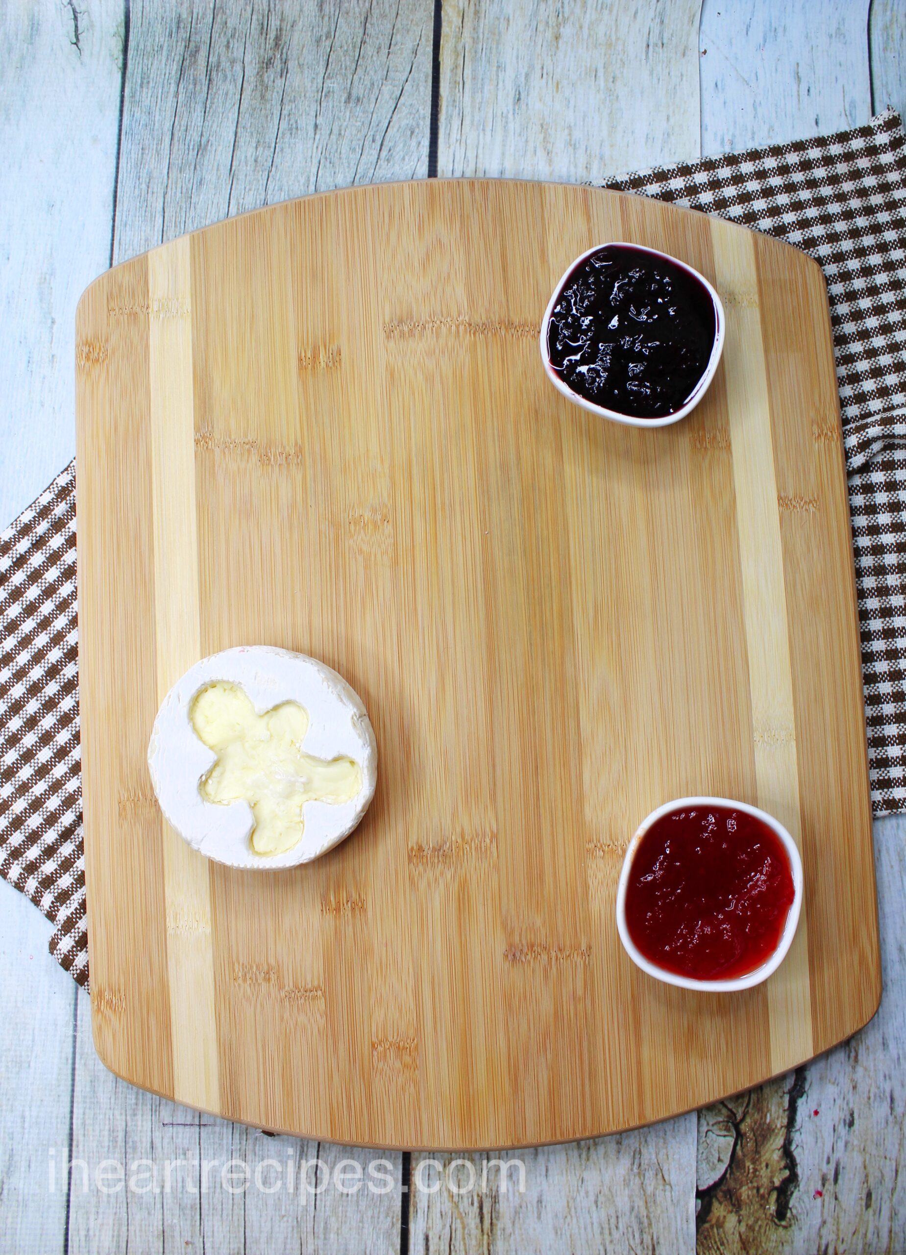 An overhead image of a wooden cutting board with two dishes of fruit jam, and one small wheel of brie cheese with a gingerbread man shape cut out of the crust of the cheese.