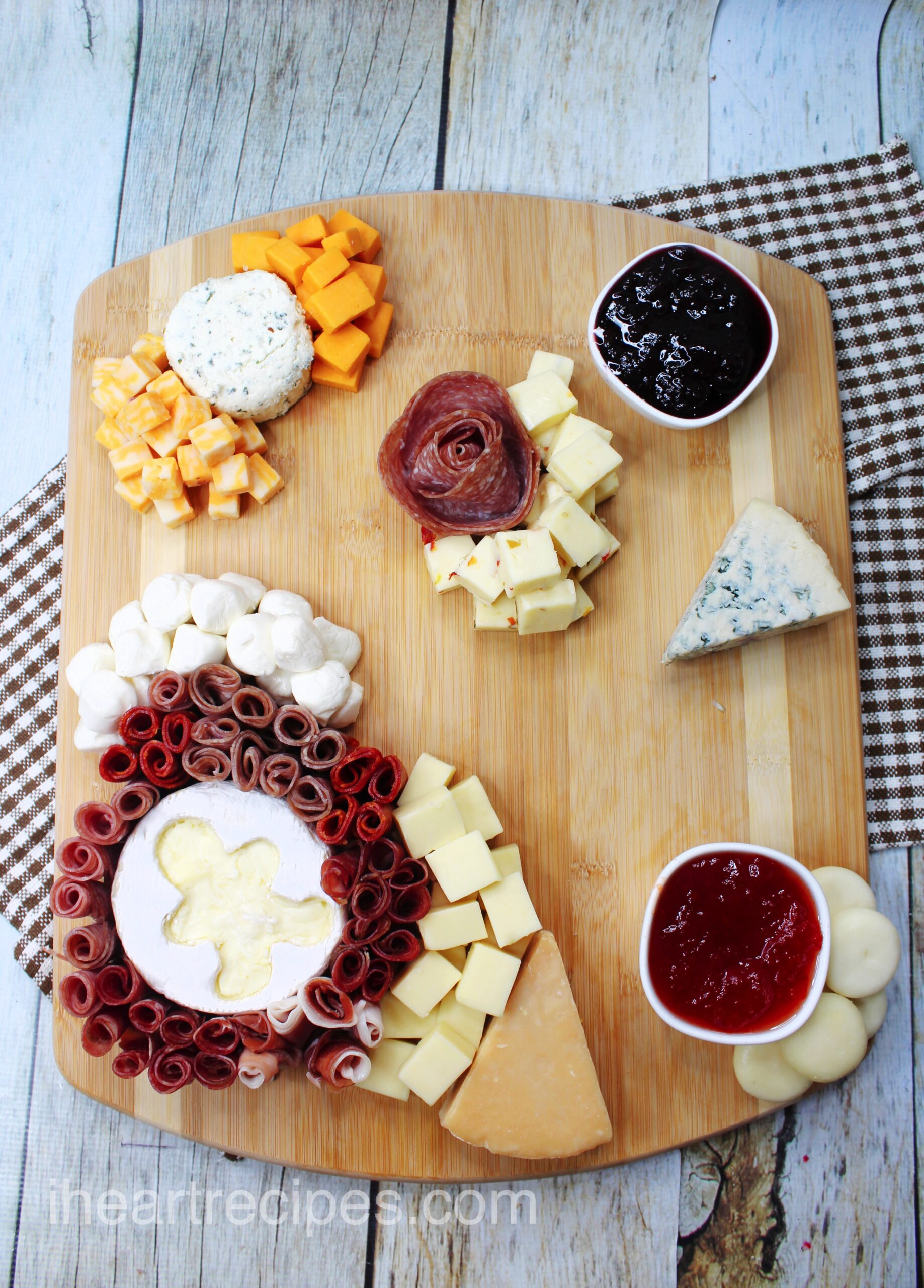 An overhead image showing the process of putting together a Christmas Charcuterie Board with cubed cheeses, jams, and rolled sliced meats arranged on a wooden cutting board.