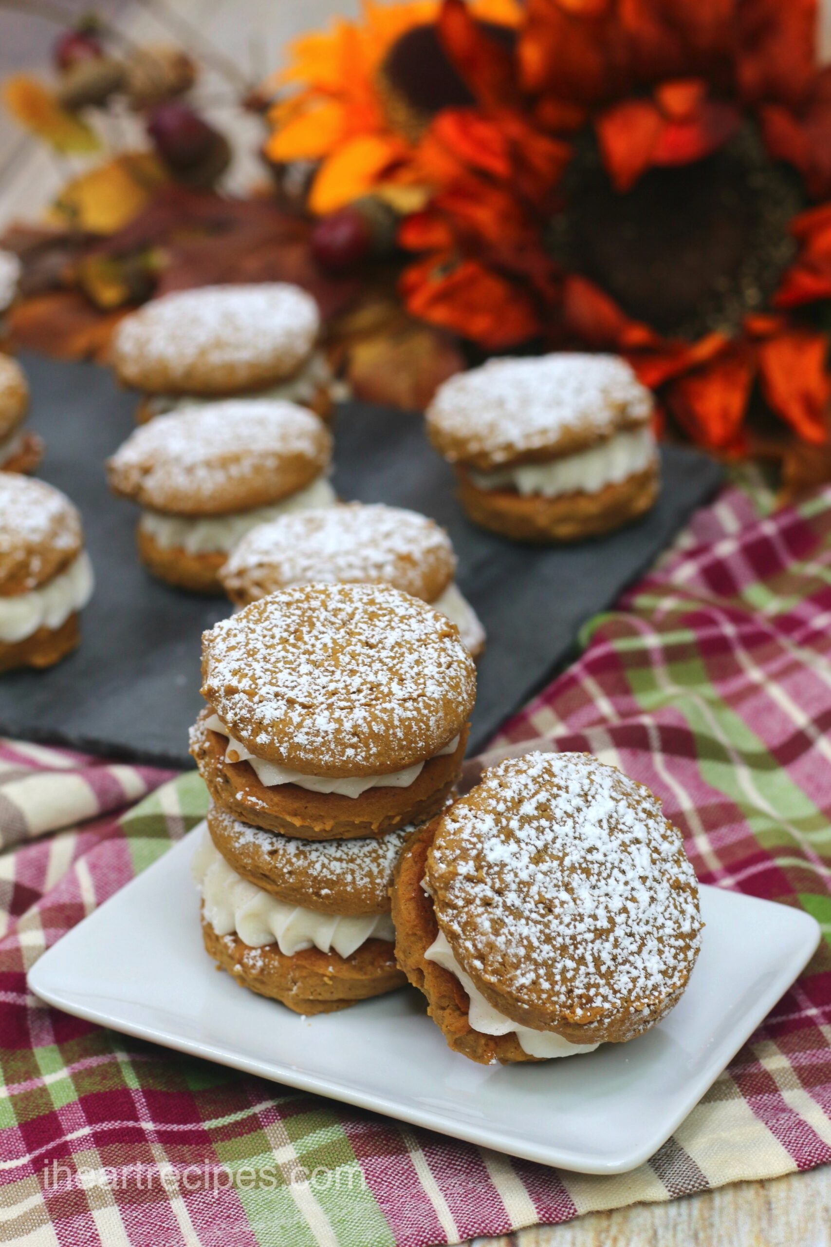 A batch of homemade pumpkin gingerbread whoopie pies sit on a platter, with three served on a small white square plate. The soft, cream-filled cookie sandwiches are dusted with powdered sugar.