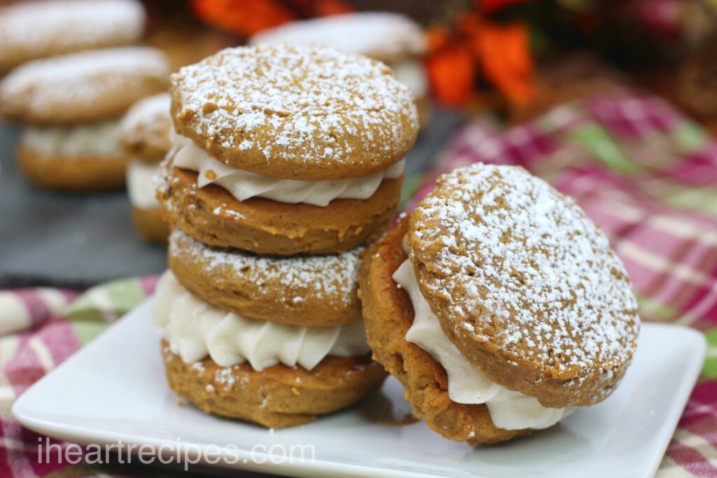 Three cream-filled pumpkin gingerbread whoopie pies are stacked together on a small plate, dusted with powdered sugar.