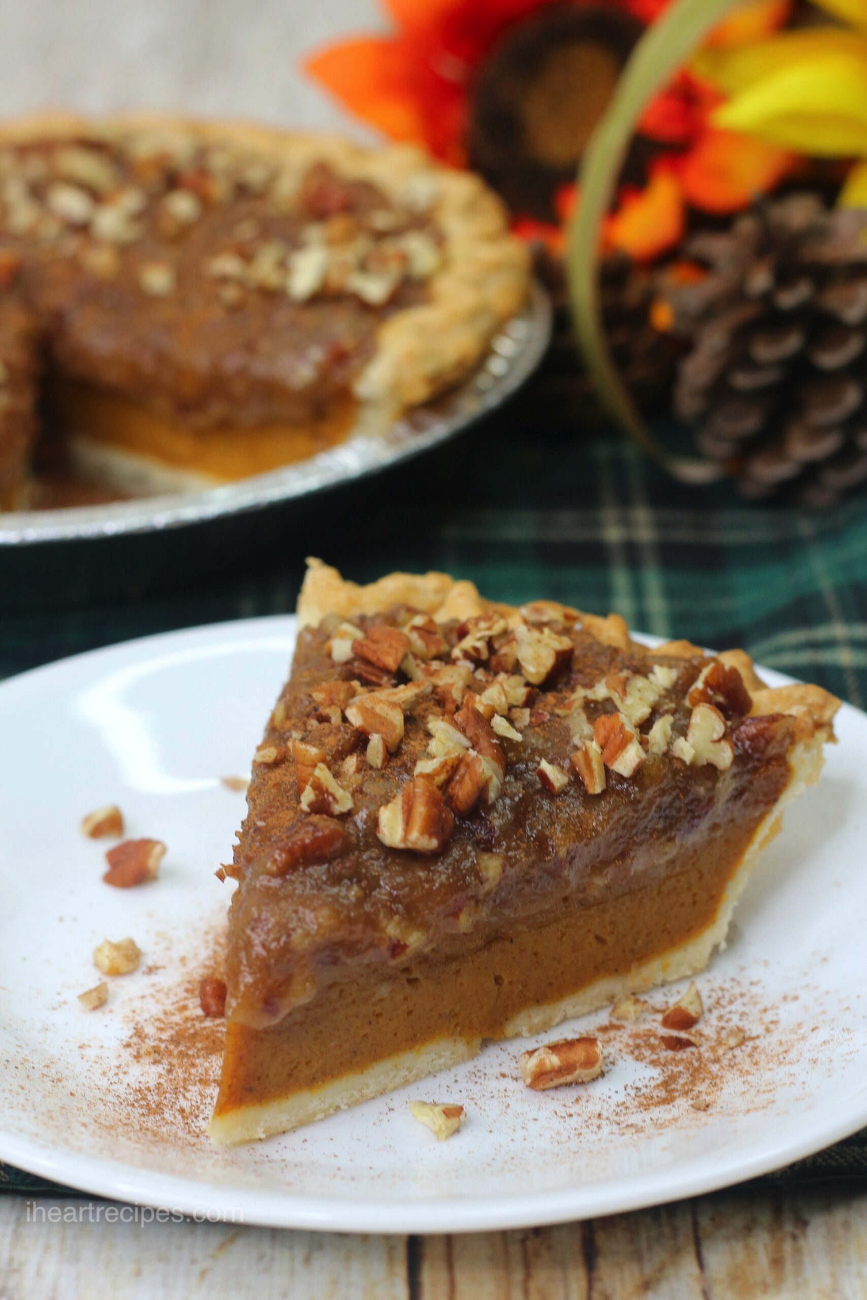 A slice of pecan pumpkin pie served on a white dessert plate. The pie is topped with chopped pecans and dusted with cinnamon. A whole pecan pumpkin pie is in an aluminum pie dish in the background.