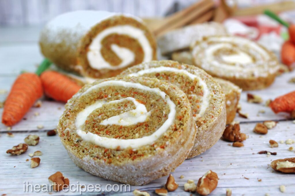 Slices of Rolled Carrot Cake stacked on a wooden table sprinkled with pecans.