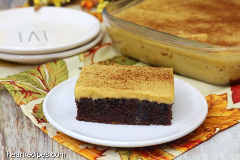 A square of chocolate Texas Sheet Cake layered with creamy pumpkin frosting and dusted with cinnamon. A glass pan of cake and a stack of white plates lay in the background.  