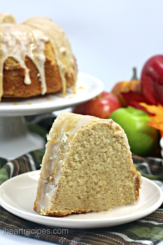 Moist caramel apple cider cake drizzled with glaze served on a white plate set on a red, green and white plaid tablecloth. The remainder of the bundt cake is set on a cake stand alongside festive autumn decorations. 