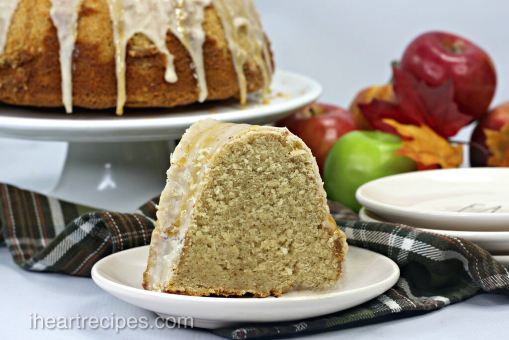 A slice of Caramel Apple Cider Cake served on a round white plate. The remainder of the bundt cake is on a stand in the background alongside red and green apples. 