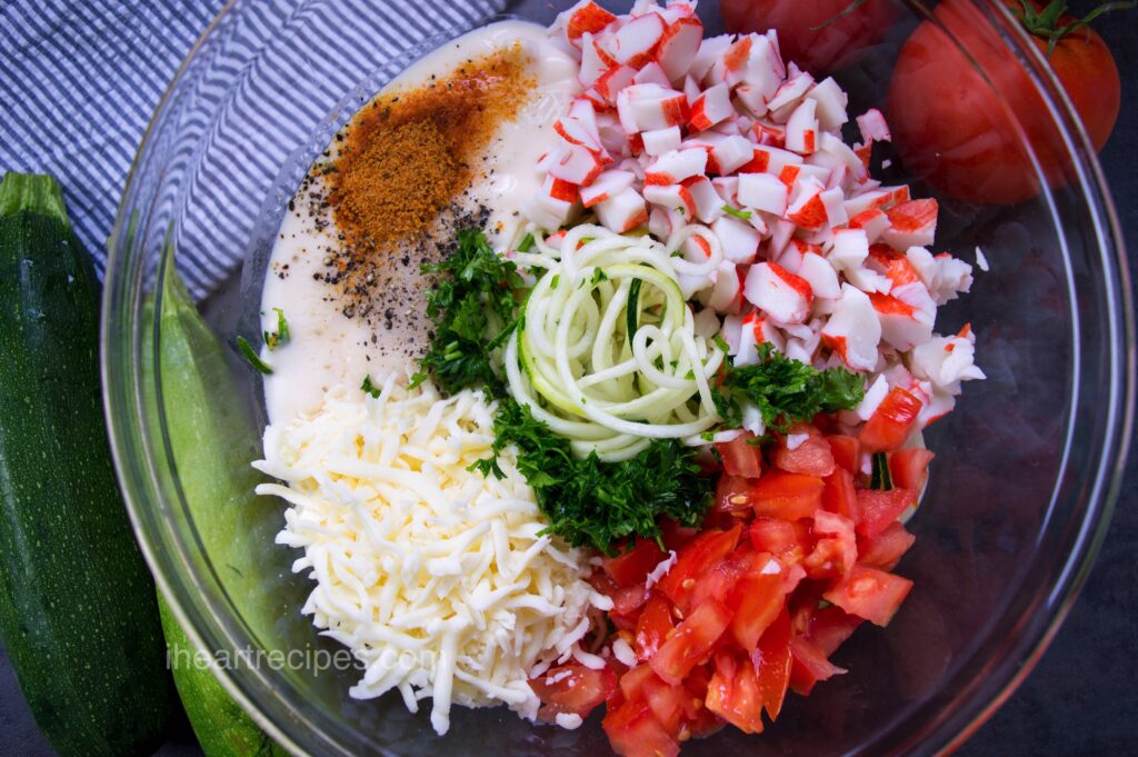 Ingredients for Zucchini and Crab Alfredo Casserole separated in a glass mixing bowl. Fresh zucchini and tomatoes lay nearby. 