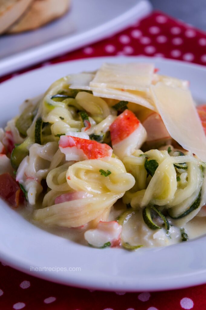 A white pasta bowl is filled with colorful vegetables and tender crab tossed in a creamy sauce and topped with shaved parmesan.