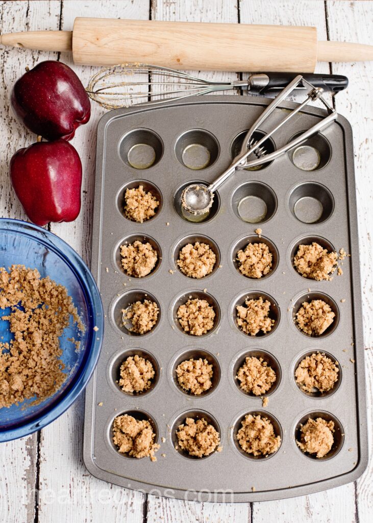 A mini muffin pan is filled with graham cracker crumbs. Baking tools lay nearby. 